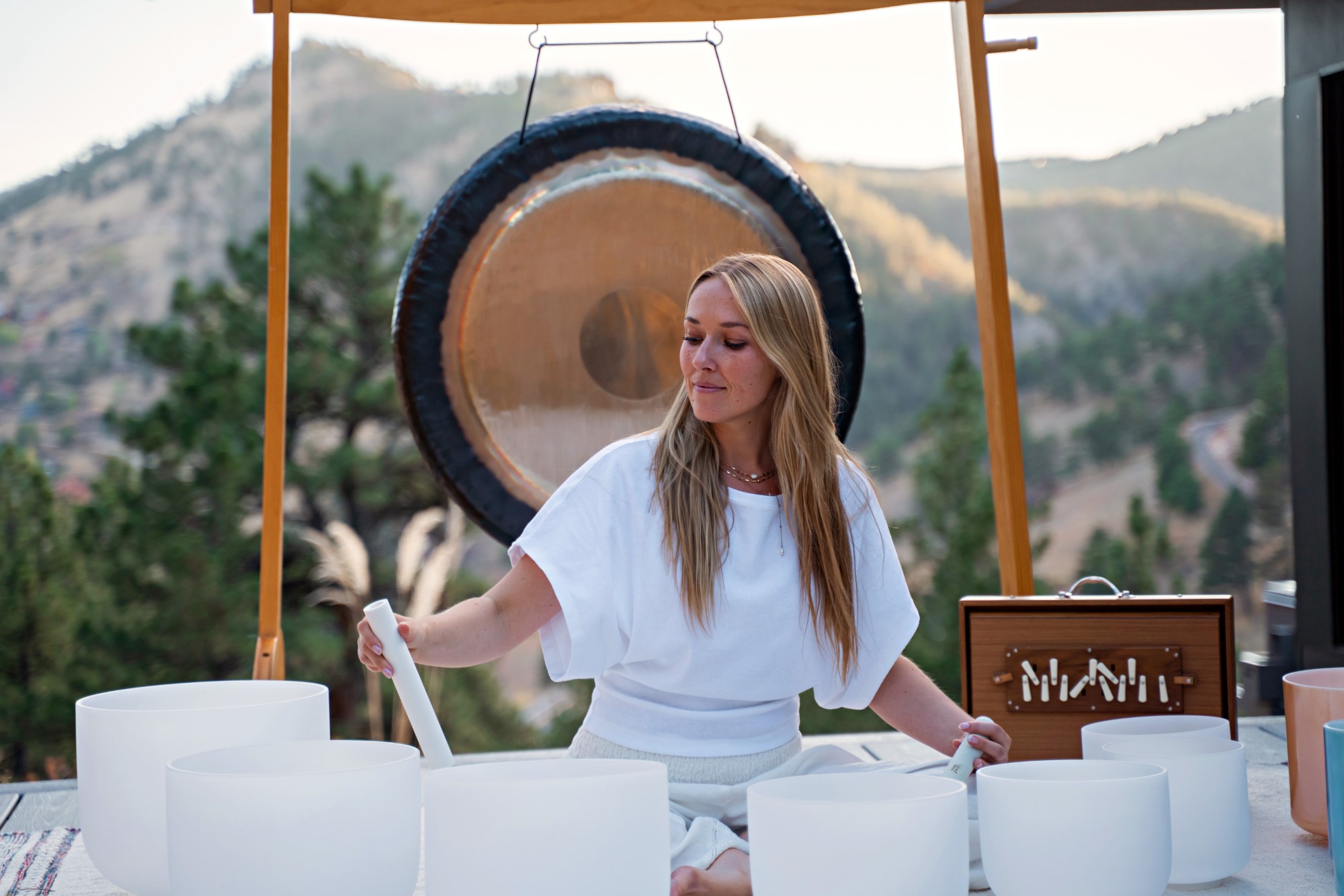 Marley Marina playing crystal singing bowls in front of a gong, with mountains behind her